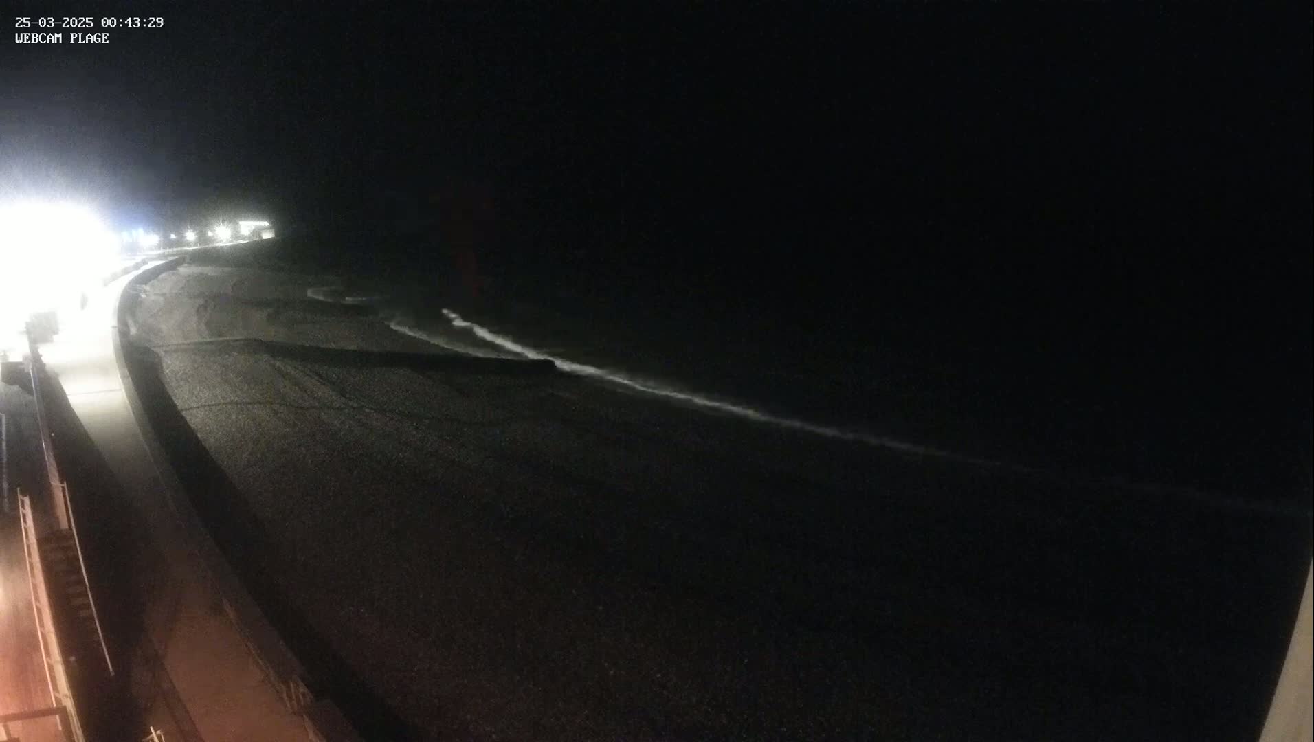 A dark, nighttime beach scene shows a shoreline with a pebble beach and a gently lapping ocean under a clear, dark sky.