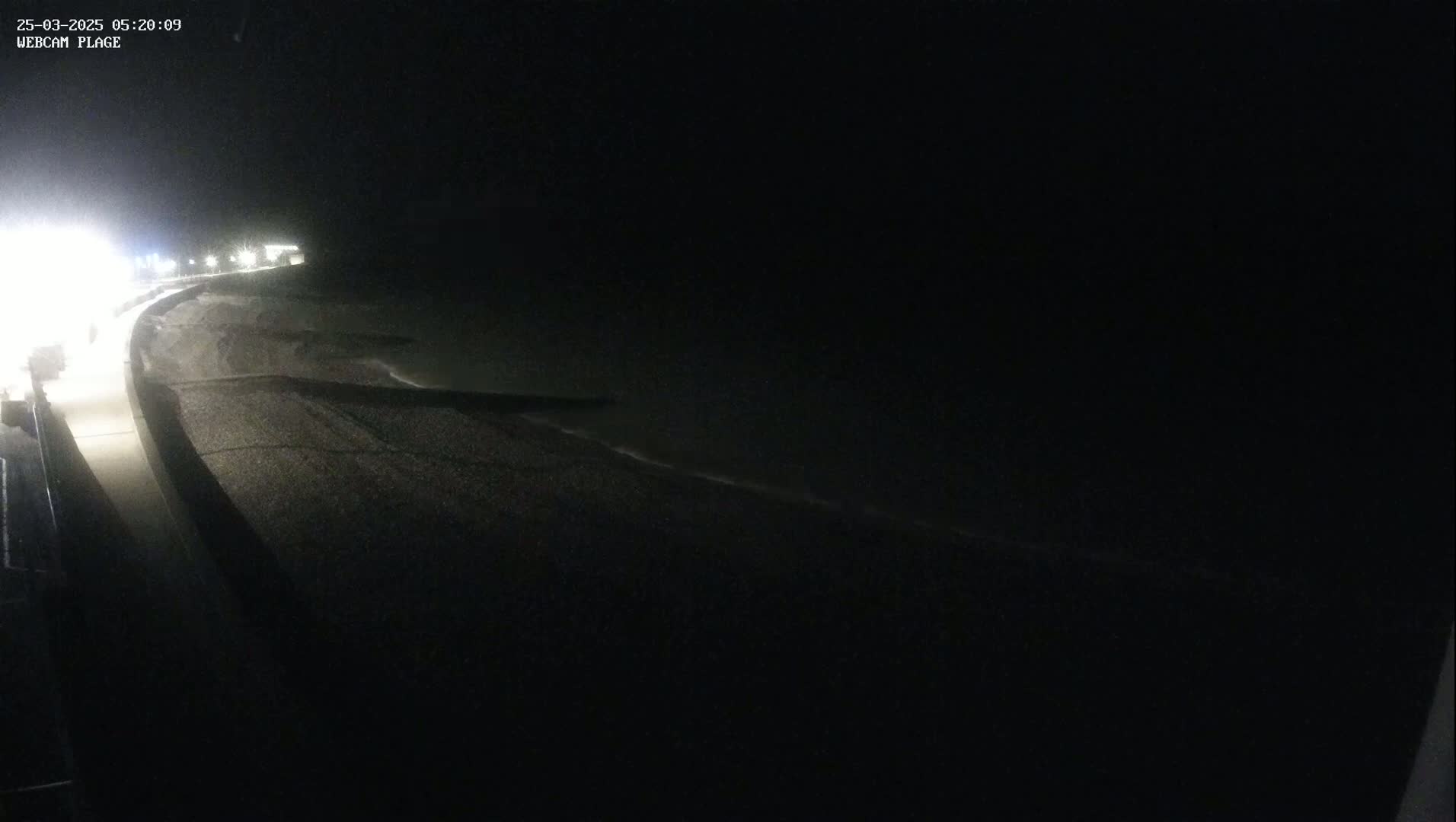 A dark, nighttime view shows a paved road next to a dark beach with gentle waves lapping the shore under a clear, black sky.