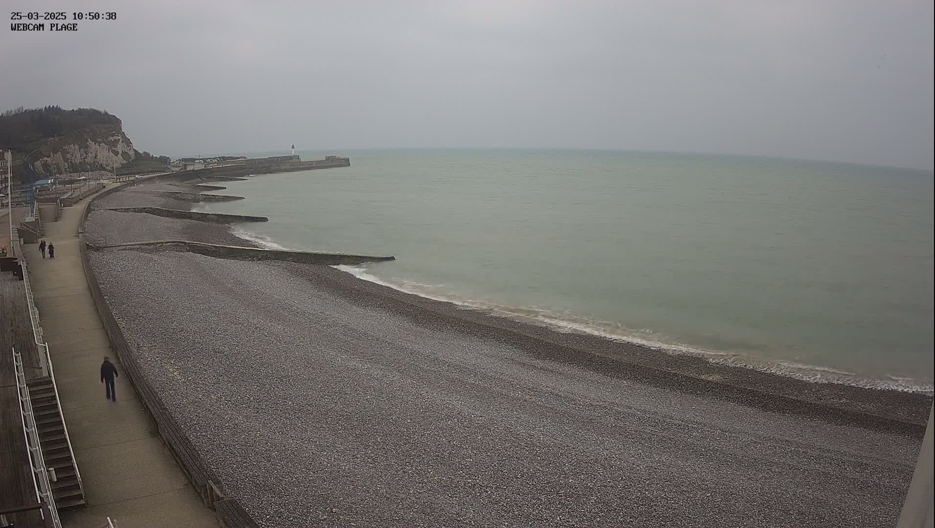 A pebble beach stretches along a calm, gray sea under an overcast sky, with a walkway and white cliffs visible in the background.