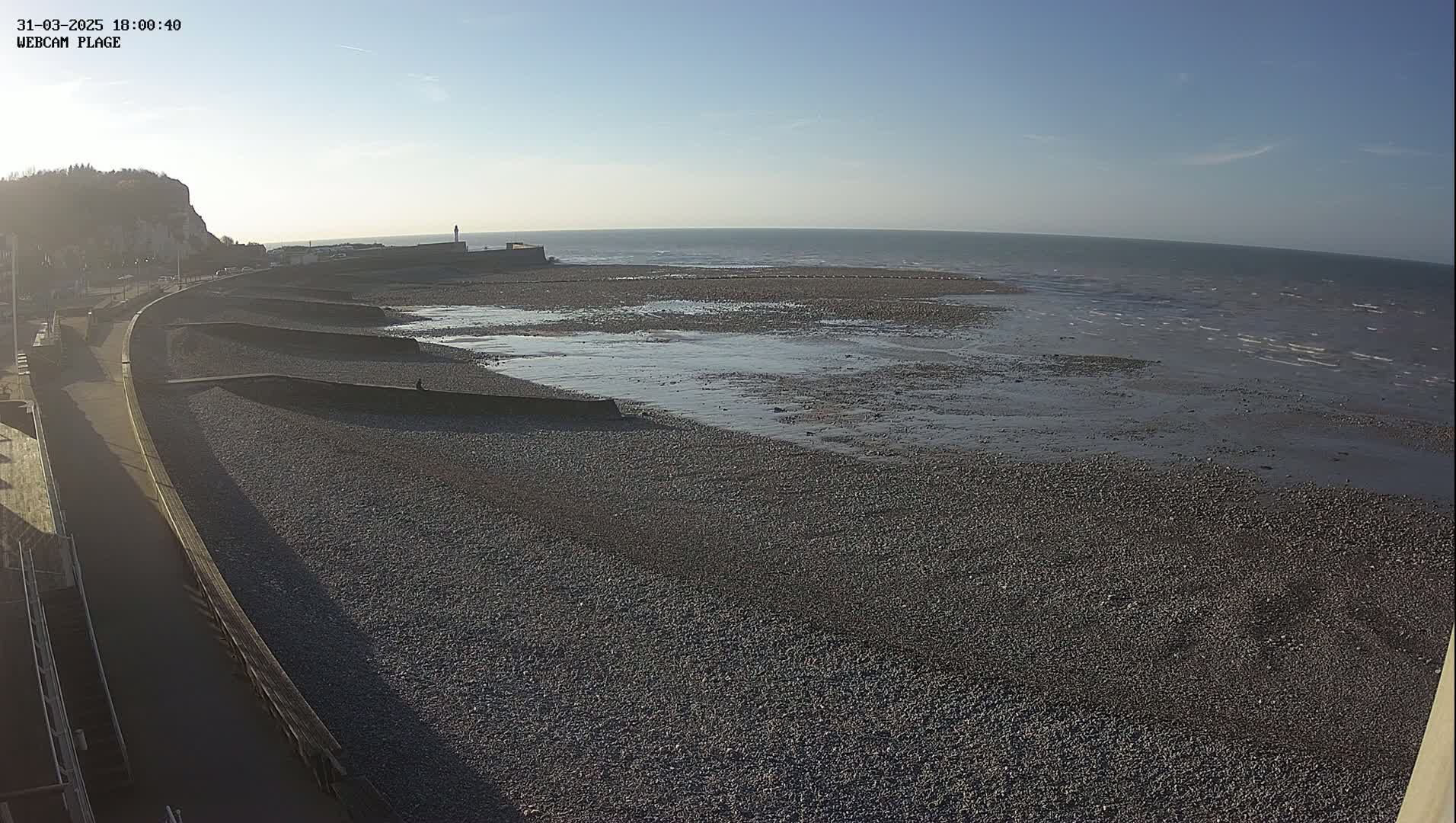 A pebble beach stretches out to a calm sea under a clear, sunny sky, with a paved walkway and sea wall alongside a cliff in the background.
