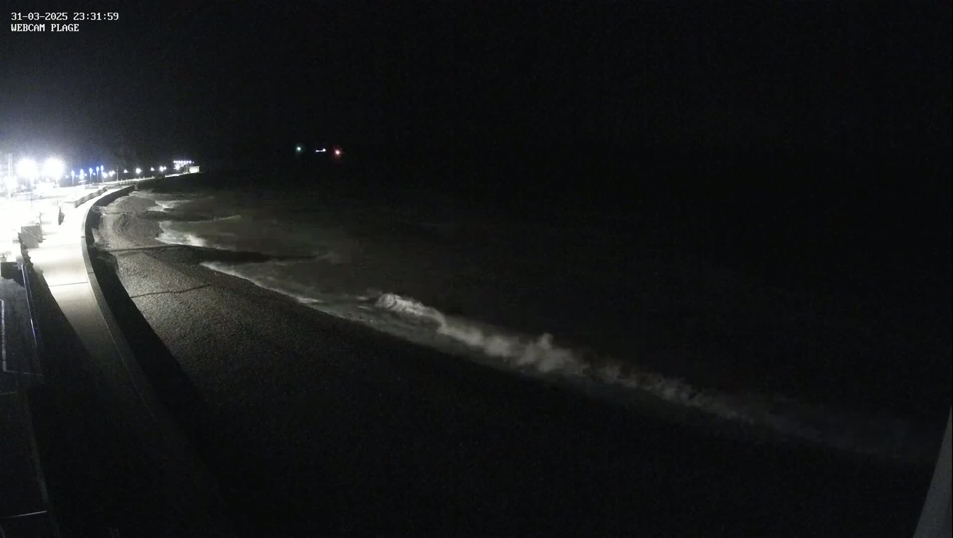 At night, waves crash on a dark, pebbled shoreline next to a lit walkway.