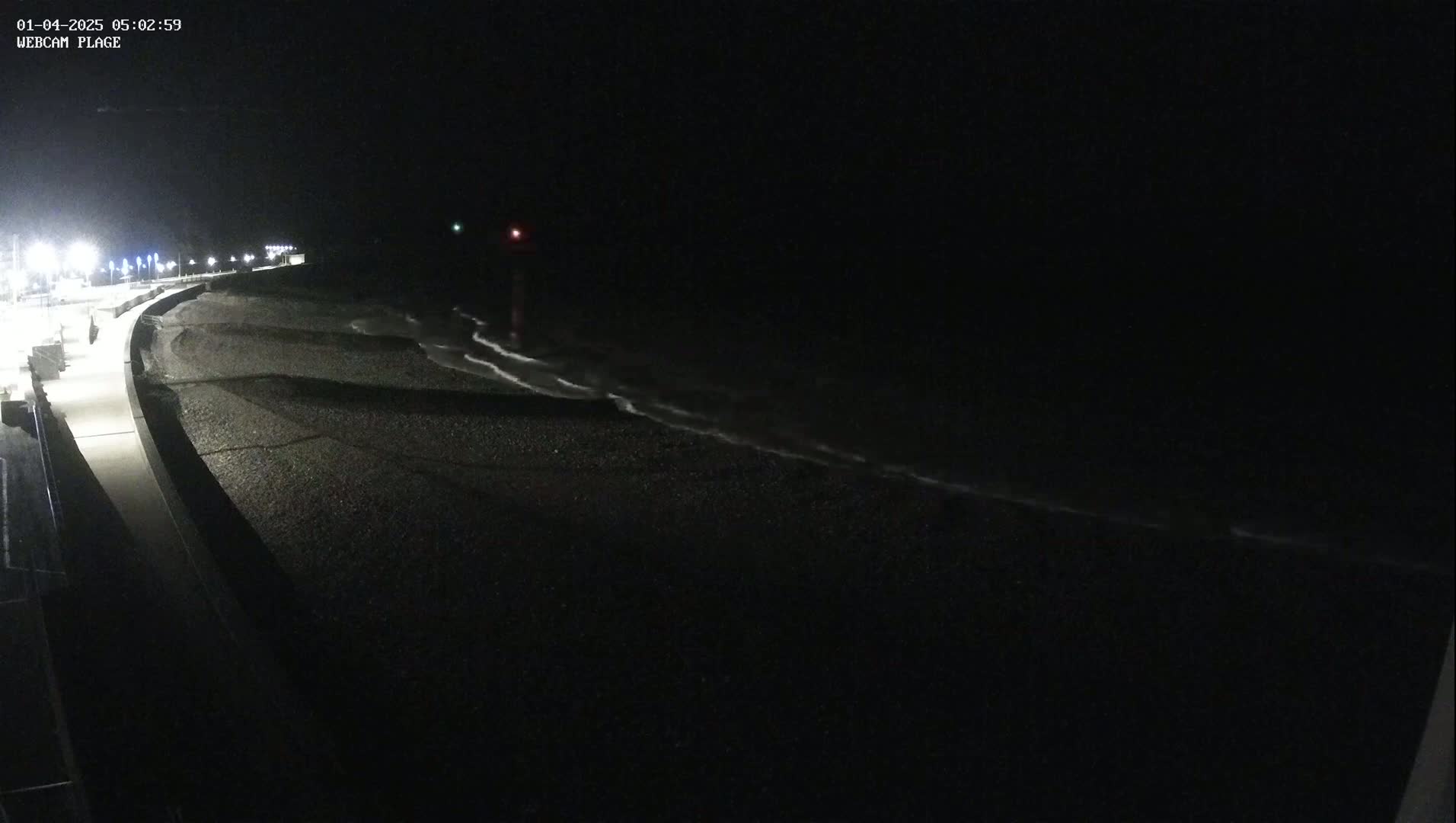 A dark, nighttime beach scene shows a paved walkway next to a pebble beach with gentle waves lapping the shore under a clear, black sky.
