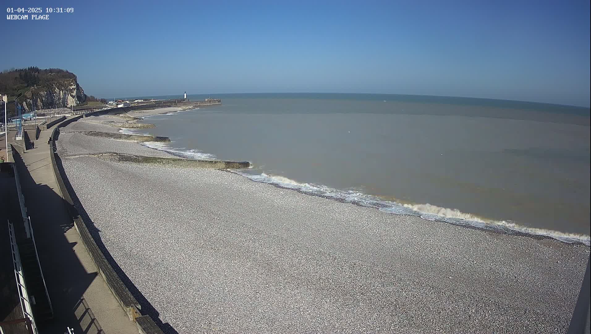 A pebble beach with small waves lapping the shore on a clear, sunny day, next to a paved walkway and a cliff in the background.