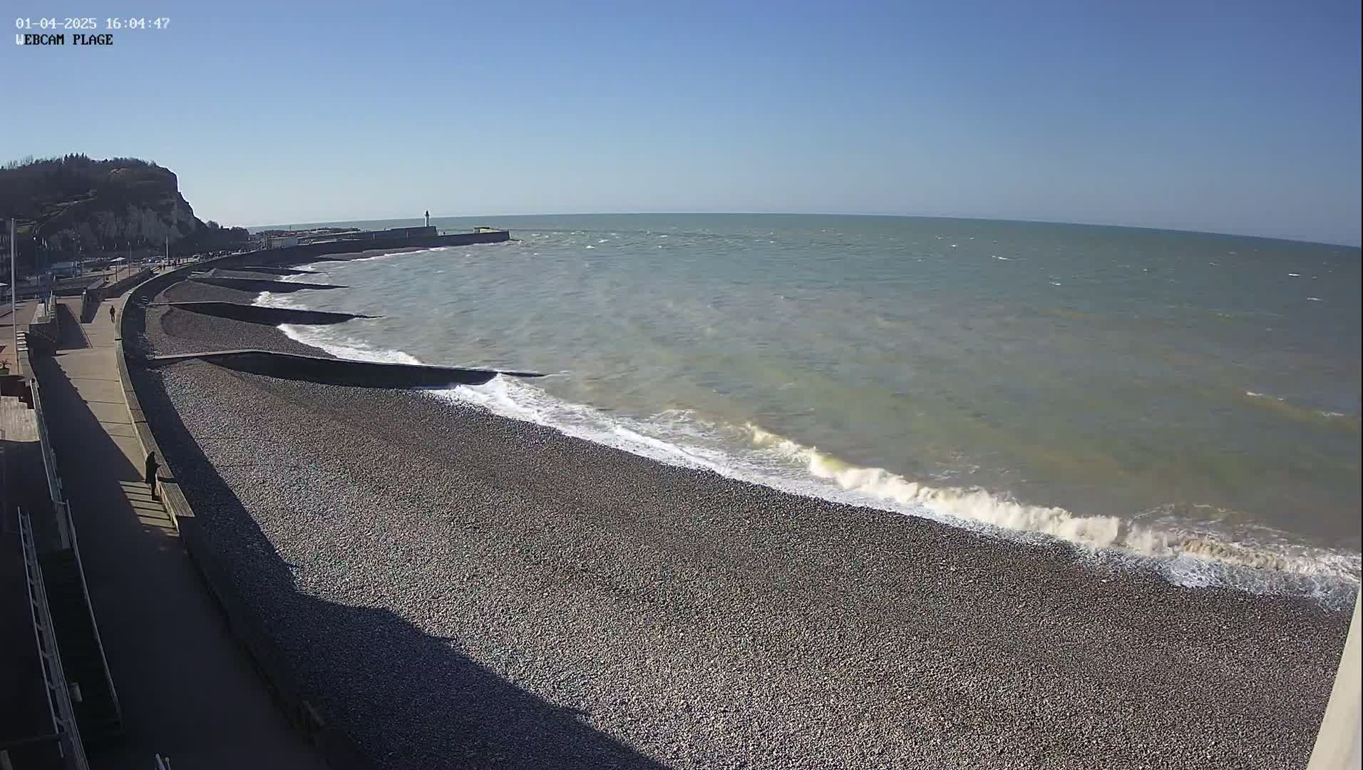 A pebble beach with gentle waves rolling onto the shore on a sunny day, backed by a paved walkway and a cliff.