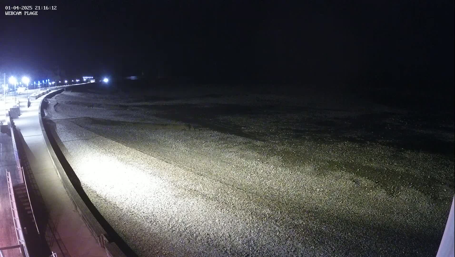 A nighttime view of a pebbled beach and a brightly lit paved walkway, with the ocean extending into a dark background.