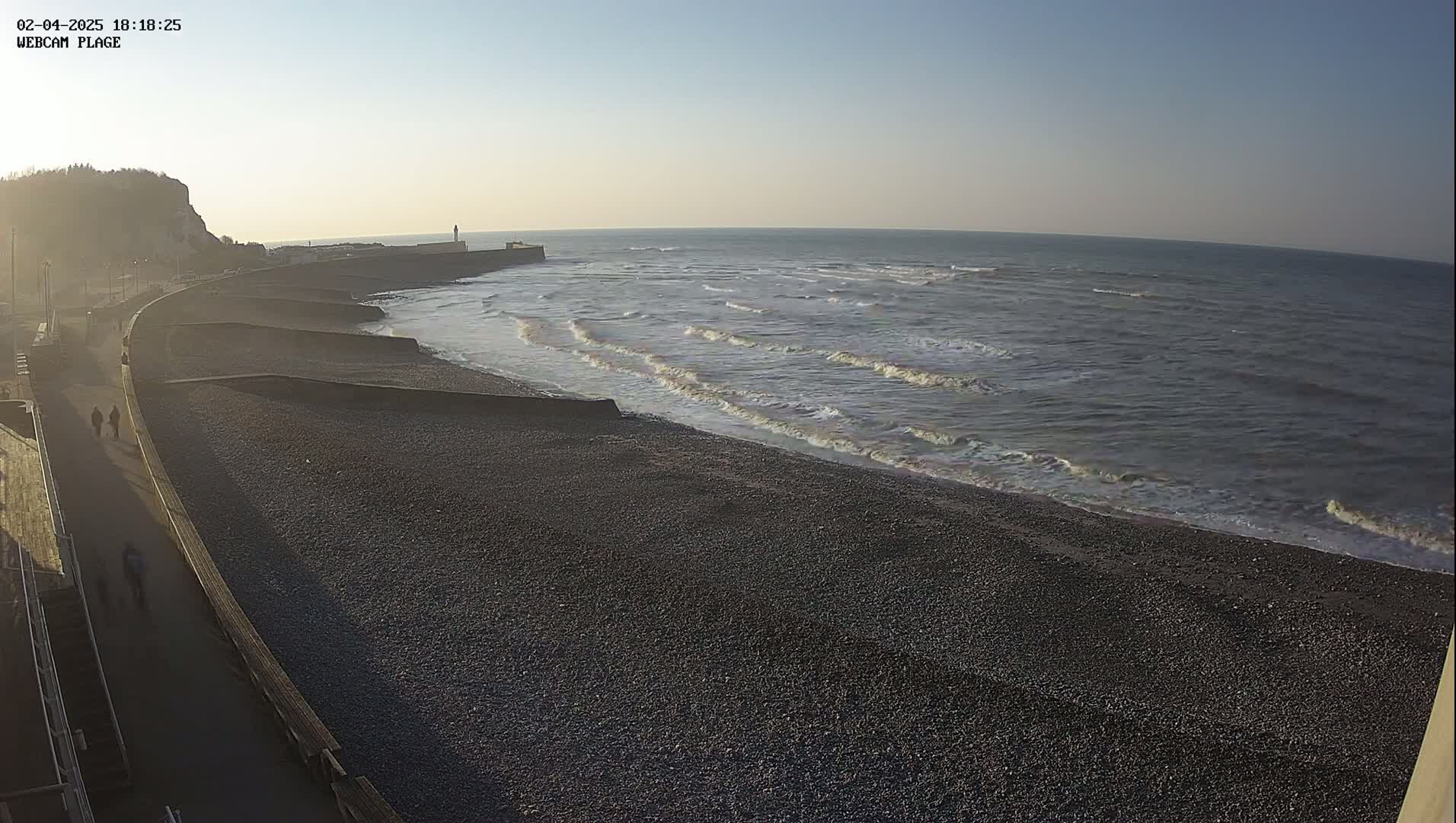 A pebble beach with a walkway and a few people walking, next to calm ocean waves under a clear sky.