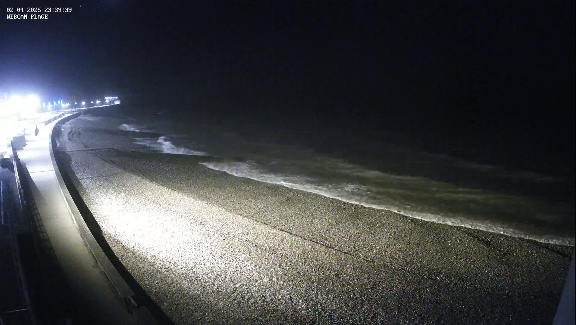 A pebble beach at night is illuminated by streetlights, with waves gently lapping the shore under a dark sky.
