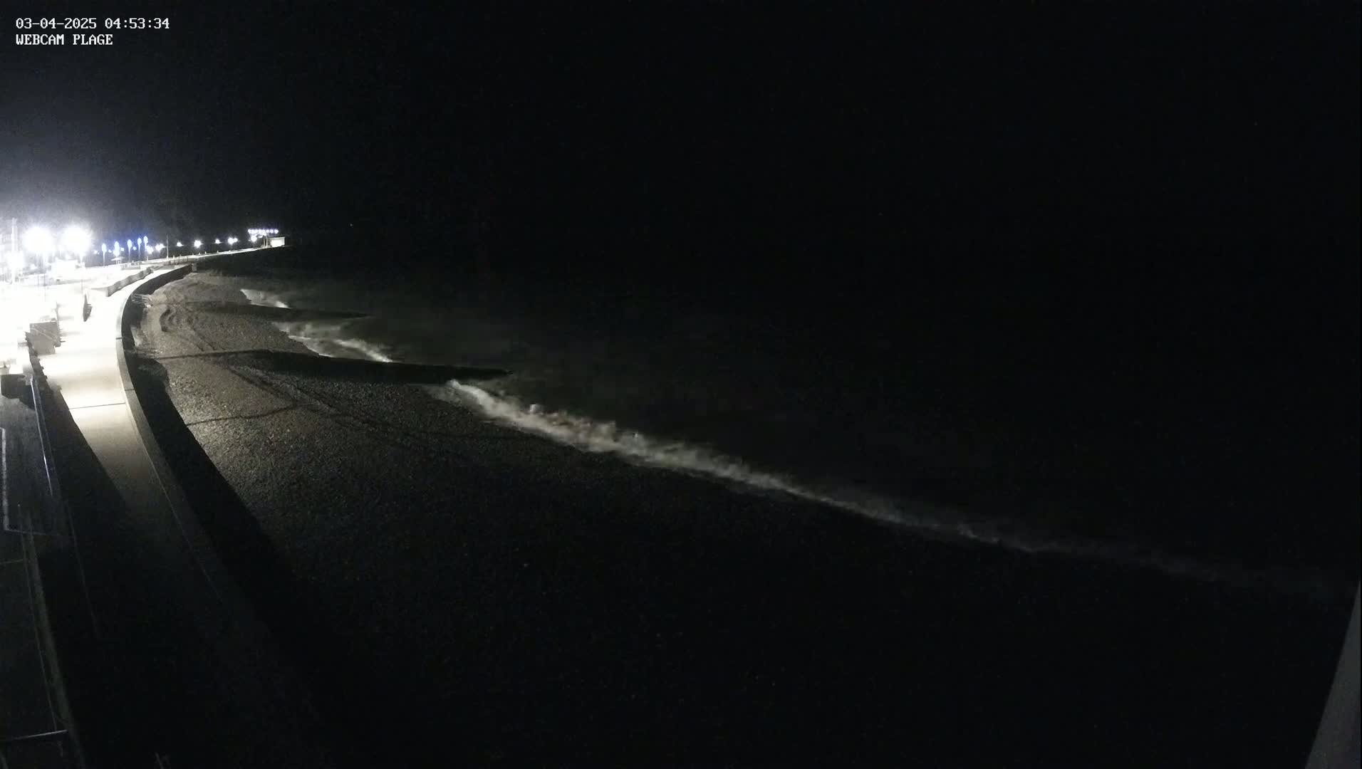 At night, waves lap a dark pebble beach next to a lit walkway.