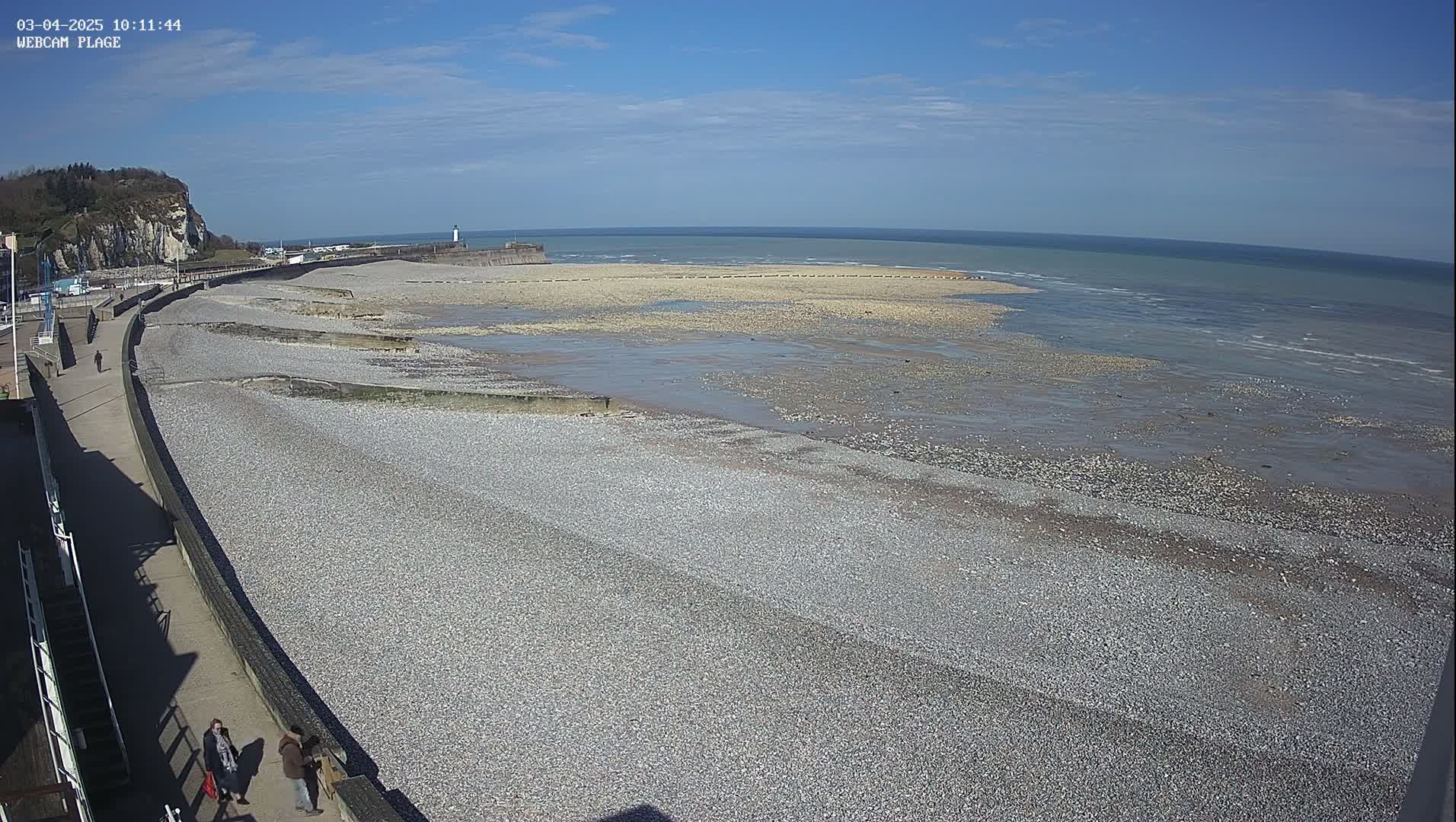 A pebble beach stretches out to a calm sea under a clear blue sky, with a few people walking along a paved walkway.