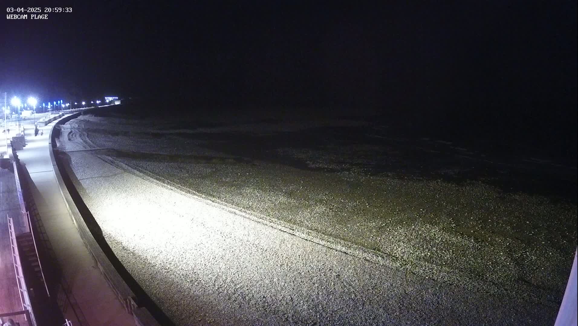 A dark, nighttime view of a pebbled beach and a paved walkway illuminated by artificial lights.