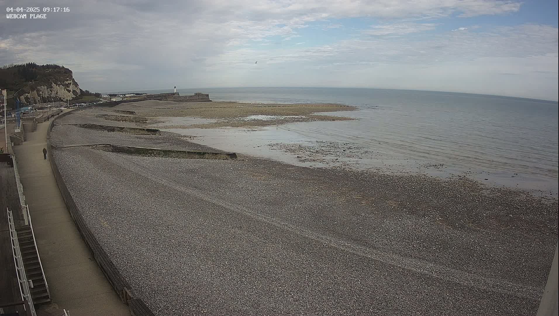 A pebble beach stretches alongside a paved walkway, leading to a calm sea under a partly cloudy sky.