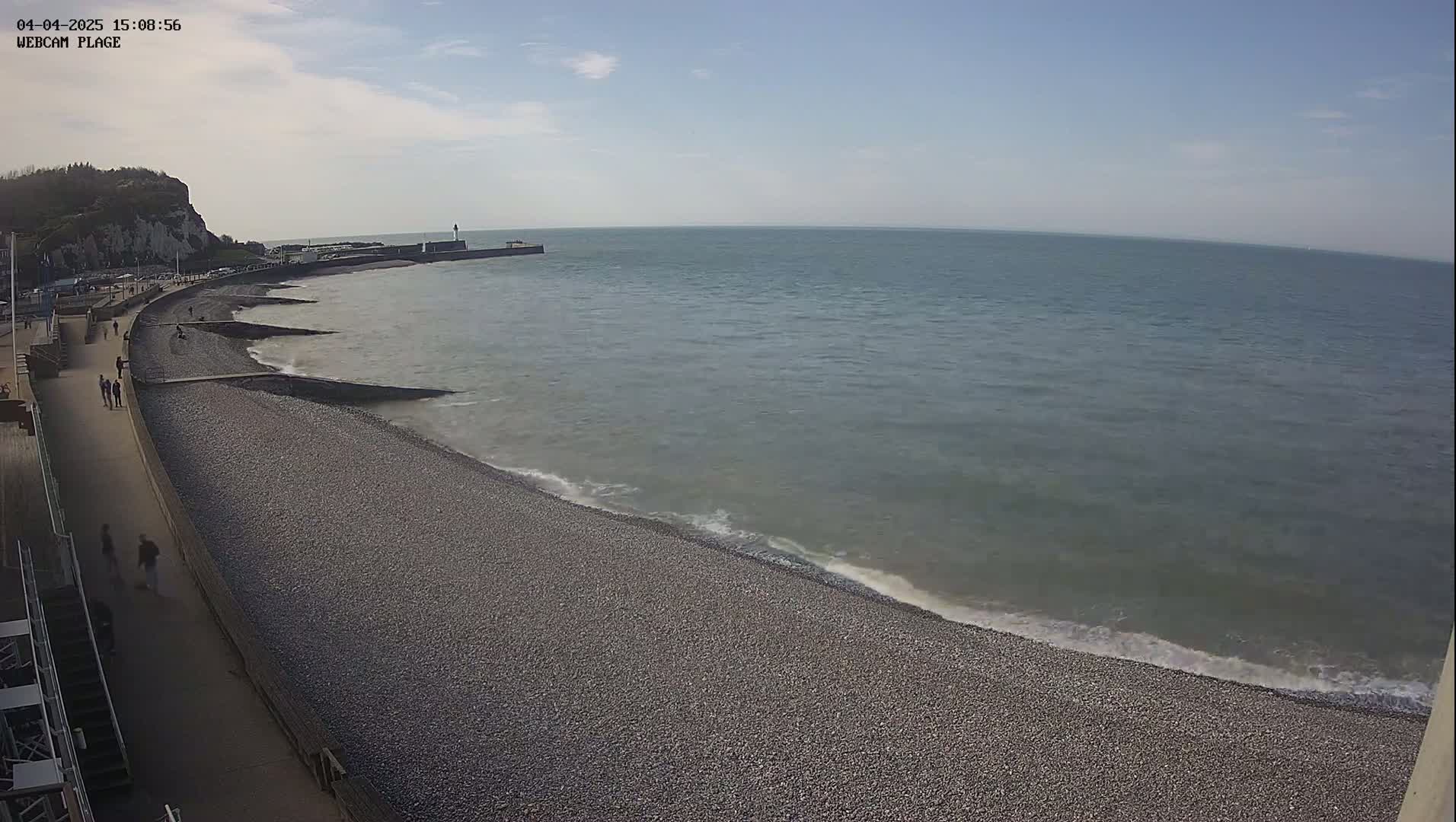 A pebble beach stretches along a calm sea under a partly cloudy sky, with people walking along a paved path beside it.