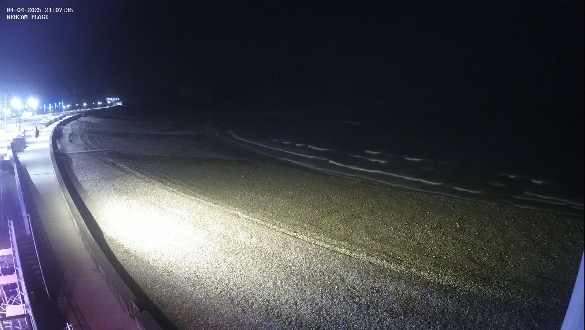 A dark, pebble beach at night is gently lapped by small waves under a clear, starless sky, with a lit walkway visible along the shore.
