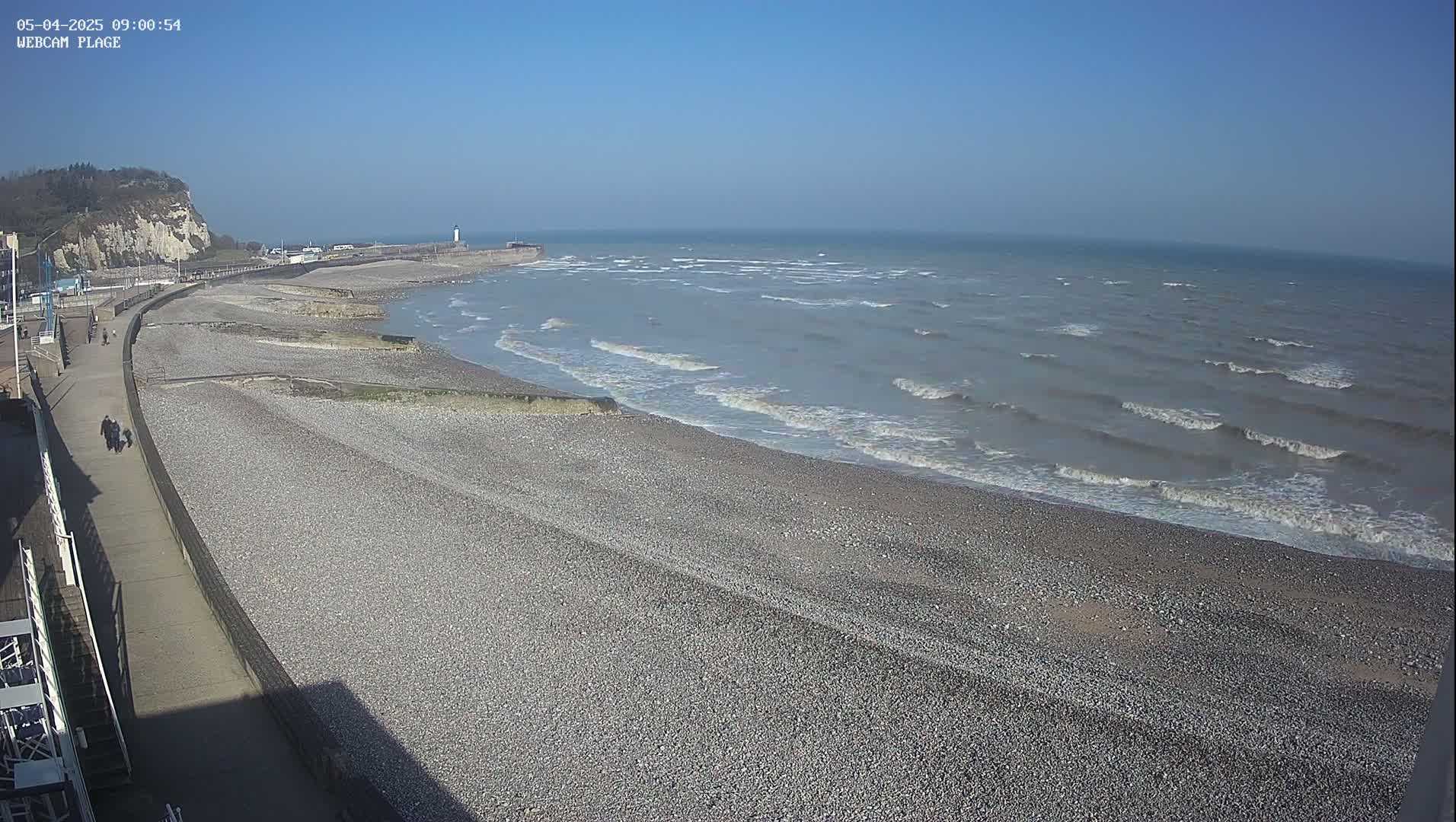 A pebble beach stretches along a coastline under a clear blue sky, with small waves lapping the shore.