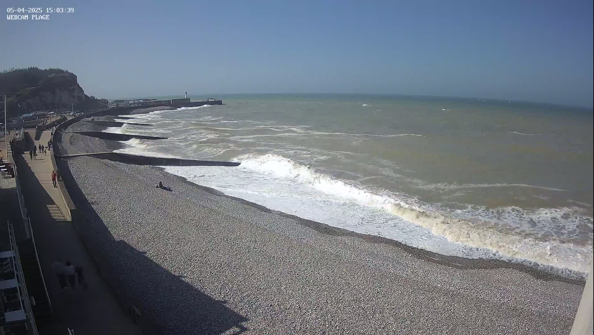 A pebble beach with a walkway and people walking along it, on a sunny day with moderate waves crashing on the shore.