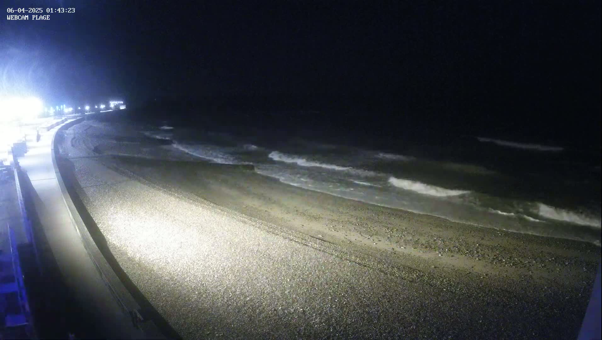 A dark, pebble beach is gently sloped toward a moderately wavy ocean at night.