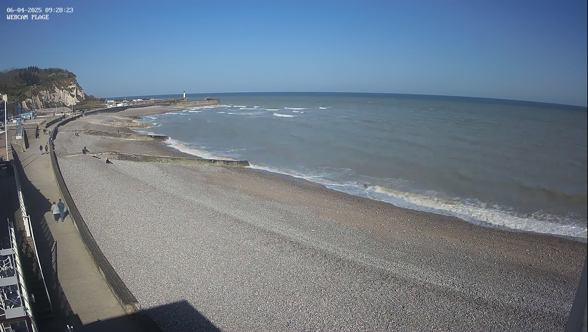 A pebble beach with a few people walking along a promenade next to a calm, grey sea under a clear blue sky.