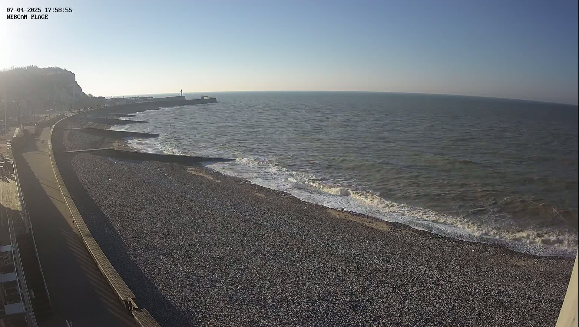 A pebble beach with gentle waves rolling onto the shore under a clear blue sky, next to a paved walkway and a cliff in the background.