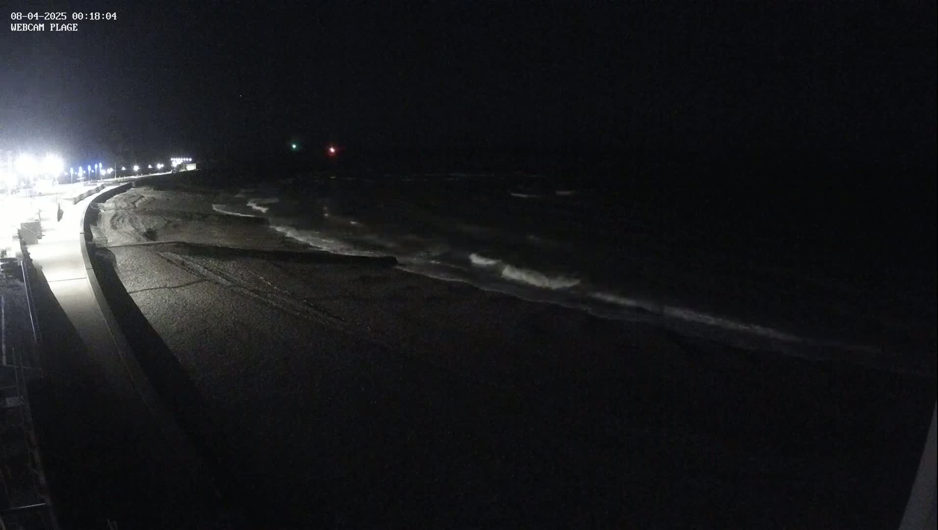 A dark, nighttime beach scene shows a paved walkway next to a dark-colored, pebble-covered shore with small waves lapping at the edge under a clear, starless sky.