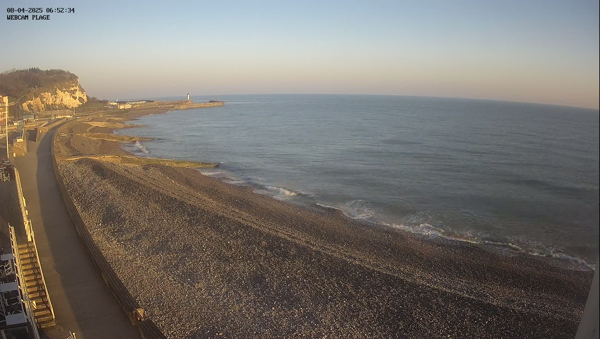 A pebble beach stretches along a calm, gray sea under a clear, pale sky, with a paved walkway and a cliff visible in the background.