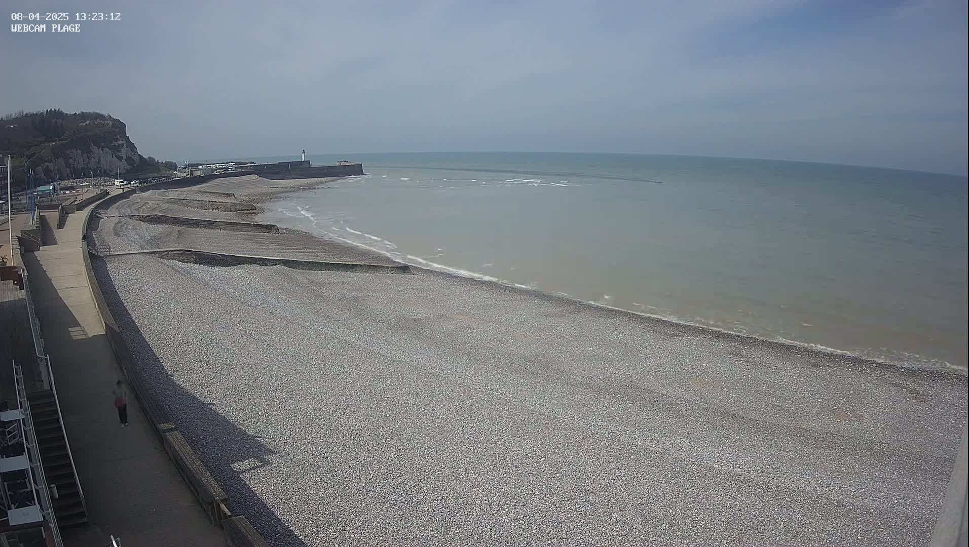 A pebble beach stretches along a calm, gray-green sea under a bright, sunny sky, with a walkway and a cliff visible in the background.
