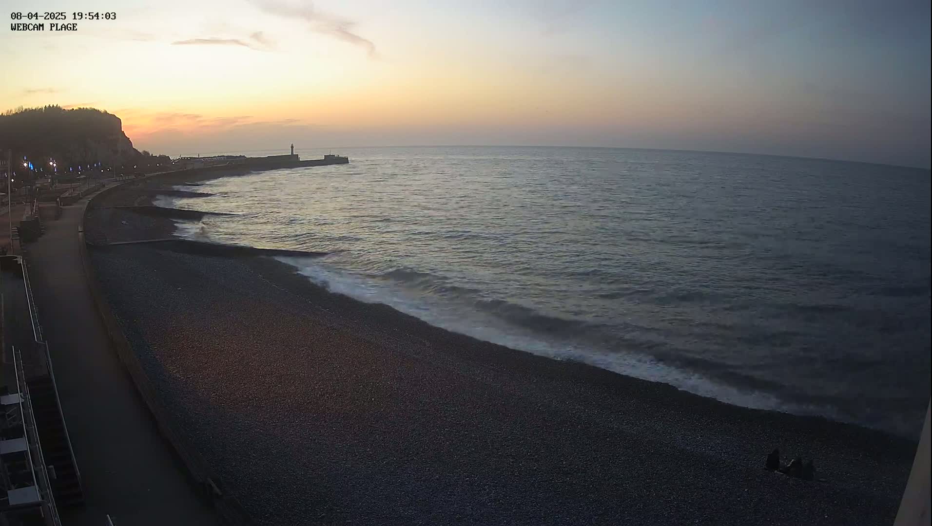 A dark pebble beach meets a calm, twilight ocean under a pale orange and dusky sky, with a small group of people sitting near the water's edge.
