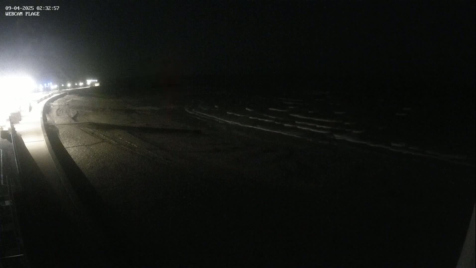 A dark, nighttime beach scene shows gently lapping waves on a mostly-dark shoreline under a clear, black sky.