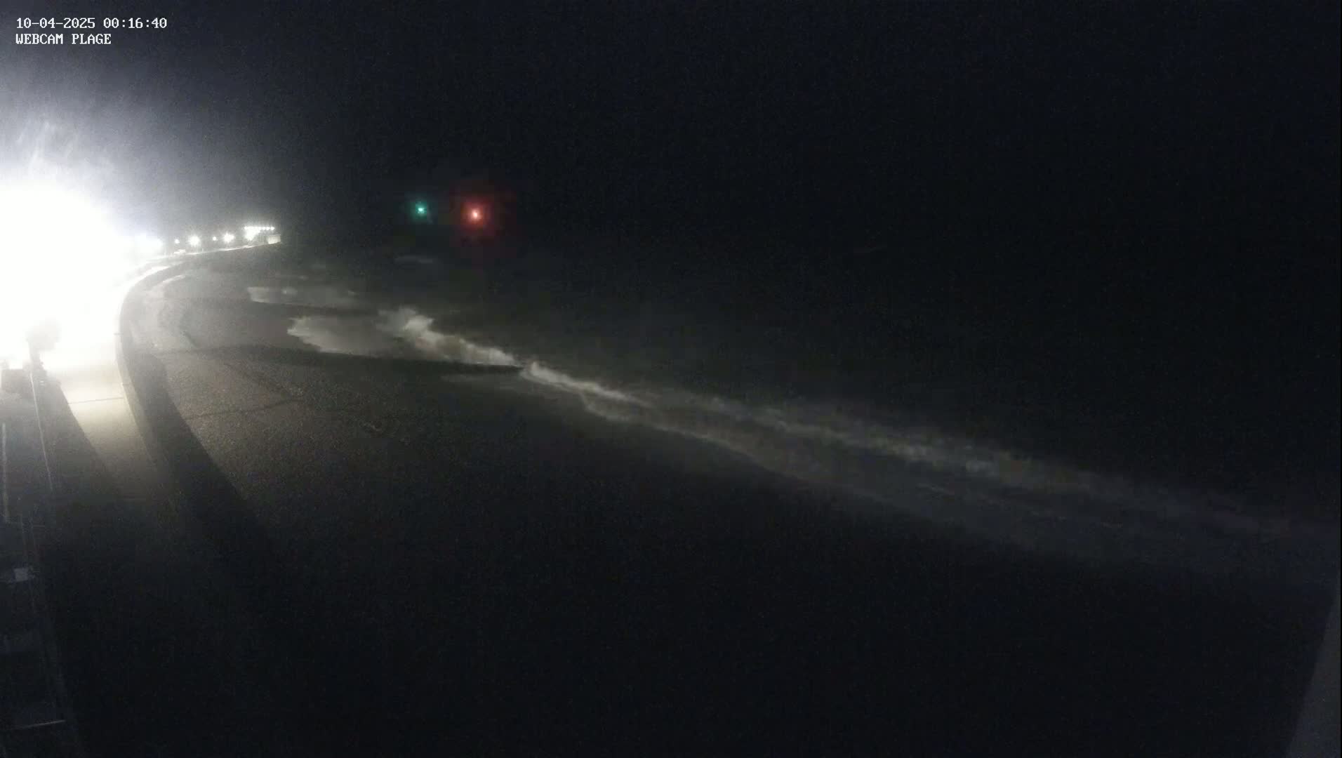 At night, waves crash on a dark shoreline next to a road with lights visible in the distance under dark, cloudy conditions.