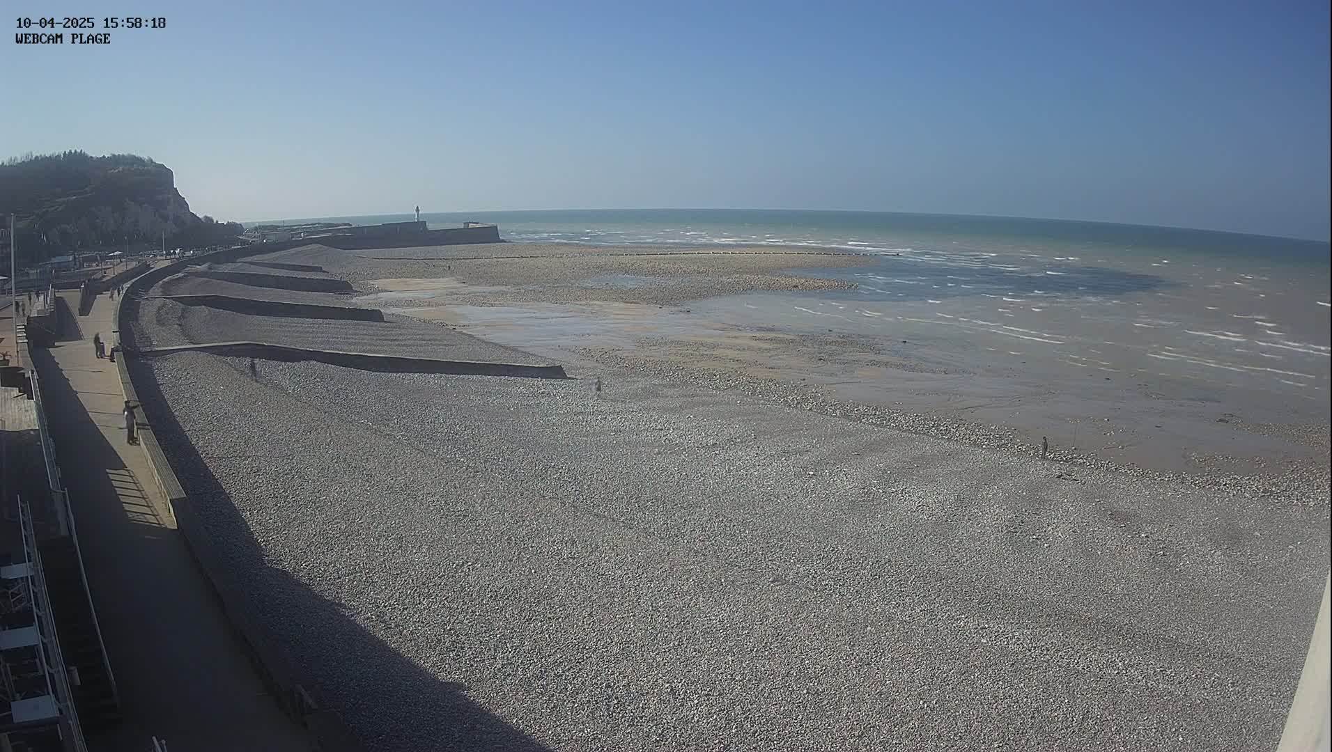 A pebble beach stretches below a paved walkway, with a calm sea under a clear blue sky.