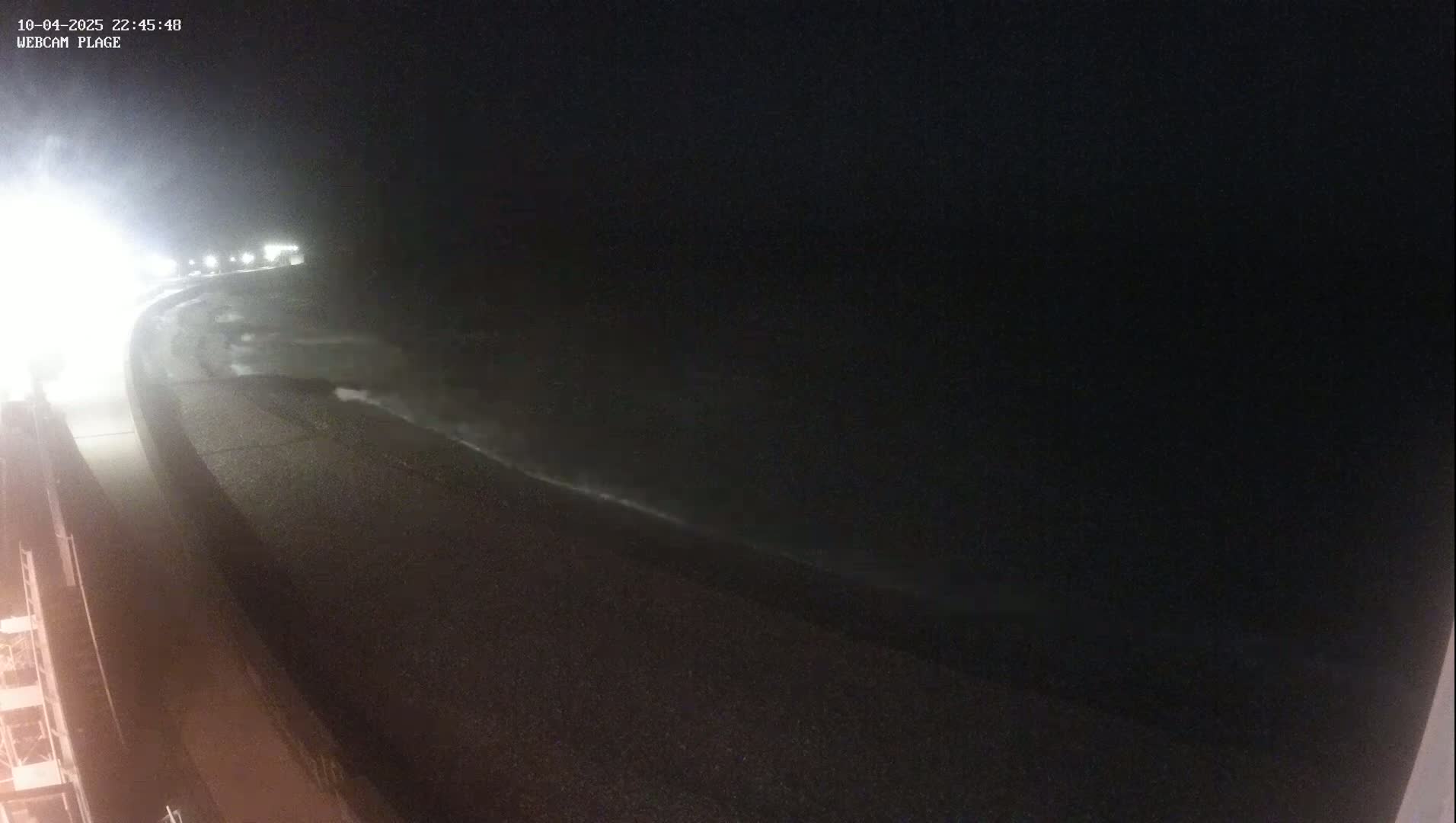 A nighttime view shows waves crashing on a dark shoreline next to a paved road under partly cloudy skies.