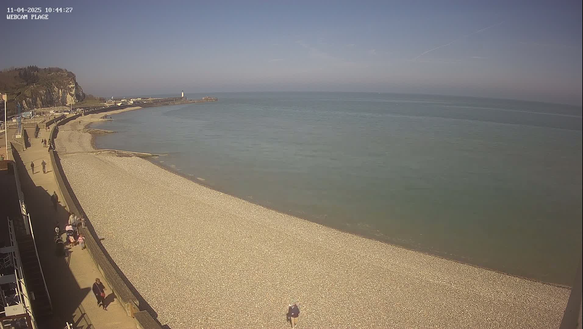 A pebble beach stretches along a calm, greenish-grey sea under a clear blue sky, with a few people walking along the shore and a walkway above.