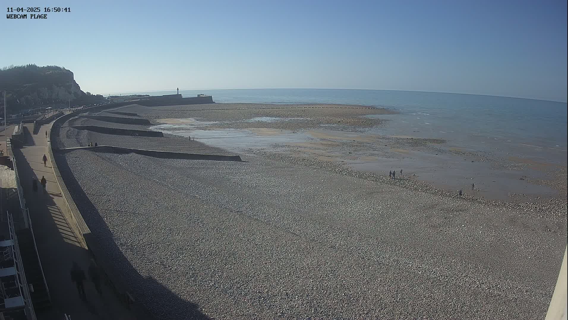 A pebble beach stretches out to a calm sea under a clear blue sky, with a few people walking on the shore and a promenade visible along the cliffside.
