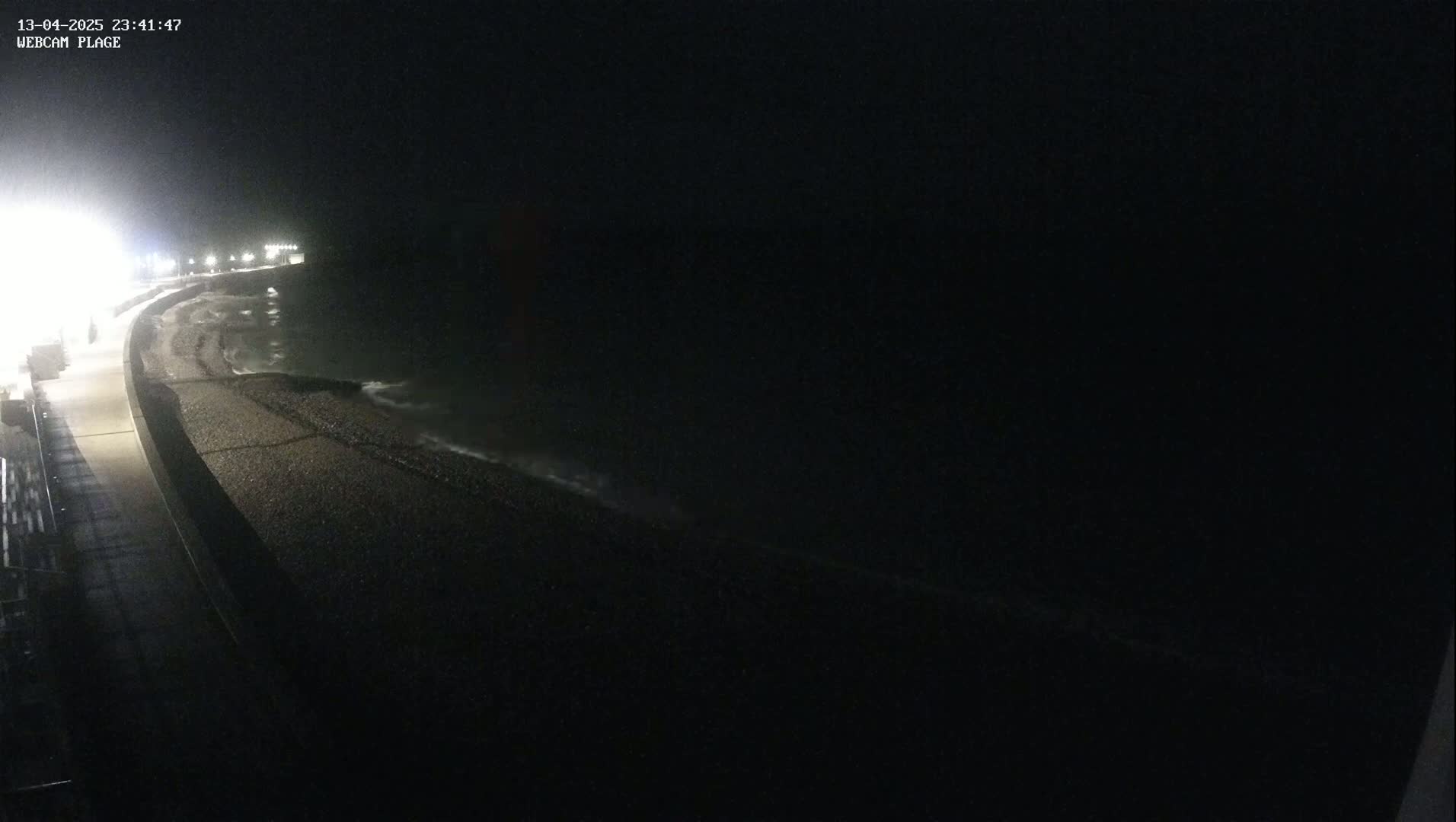 A dark, nighttime view of a pebble beach and a paved walkway along the coast, with gentle waves lapping the shore.