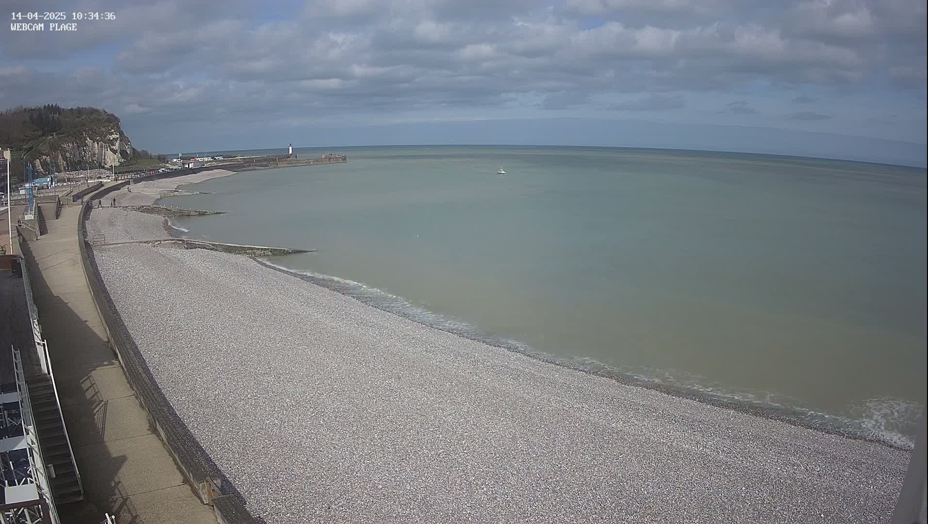 A pebble beach stretches along a calm, light-grey sea under a partly cloudy sky, with a walkway and a distant cliff visible.