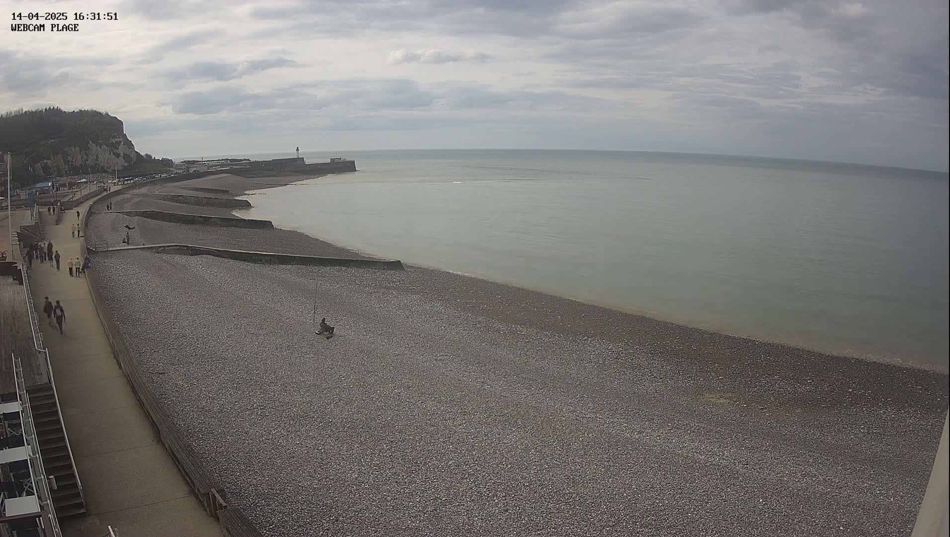 A pebble beach stretches along a calm, gray sea under an overcast sky, with a walkway and a few people visible along the shoreline.