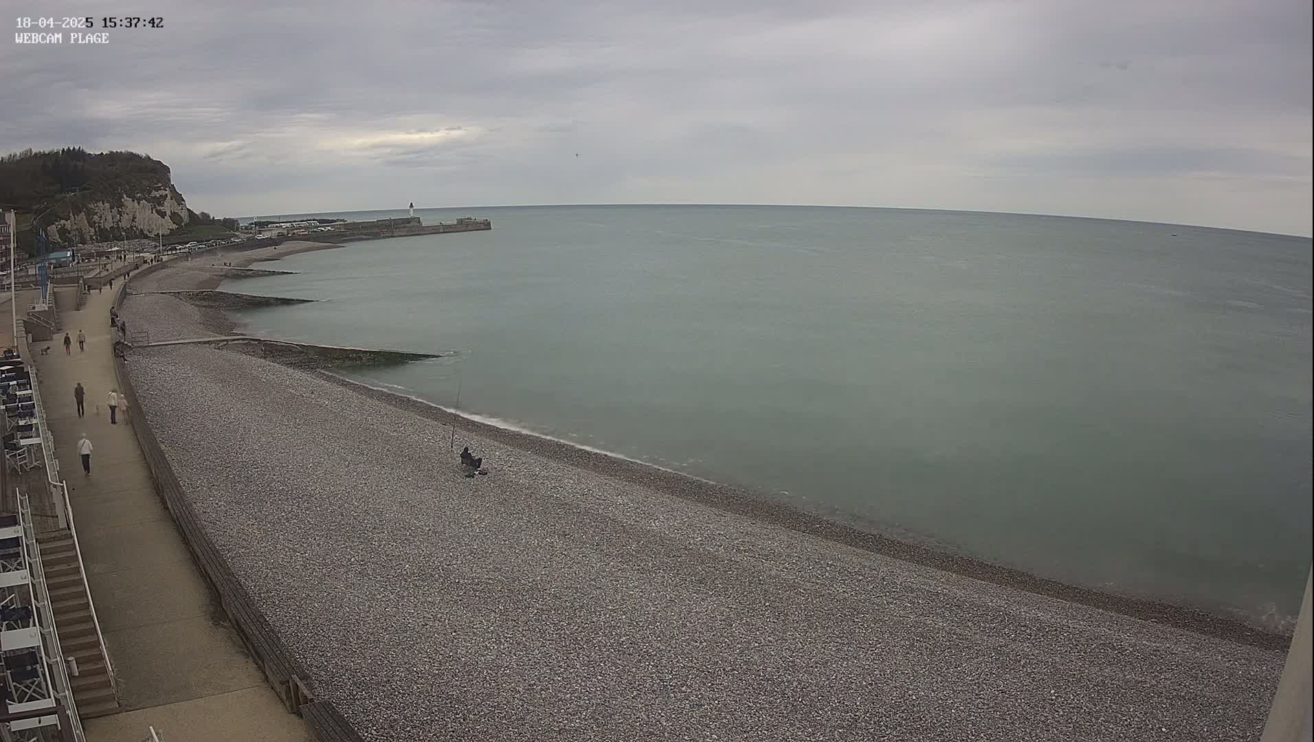 A pebble beach curves alongside a paved walkway, leading to a calm, gray sea under an overcast sky.