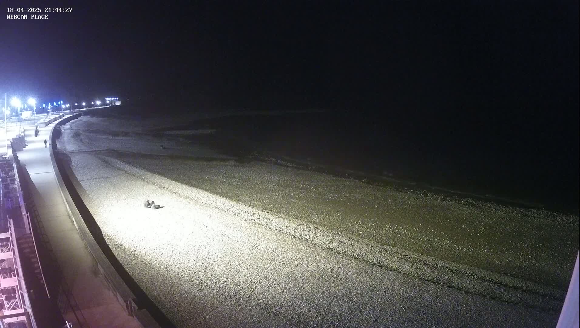 A dark, nighttime view of a pebbled beach and adjacent walkway, lit by streetlights, with a calm, dark body of water beyond.