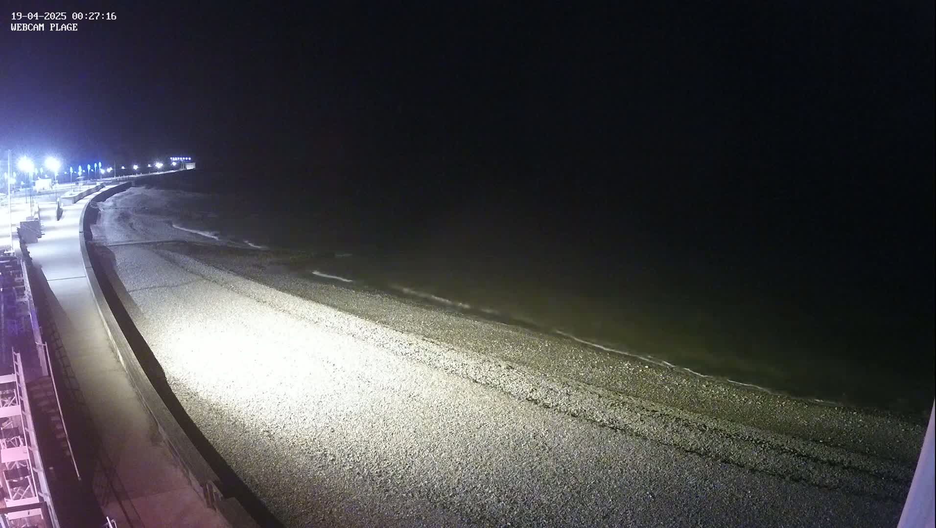 A dark, nighttime view of a pebble beach with gentle waves lapping the shore, illuminated by lights along a paved walkway.