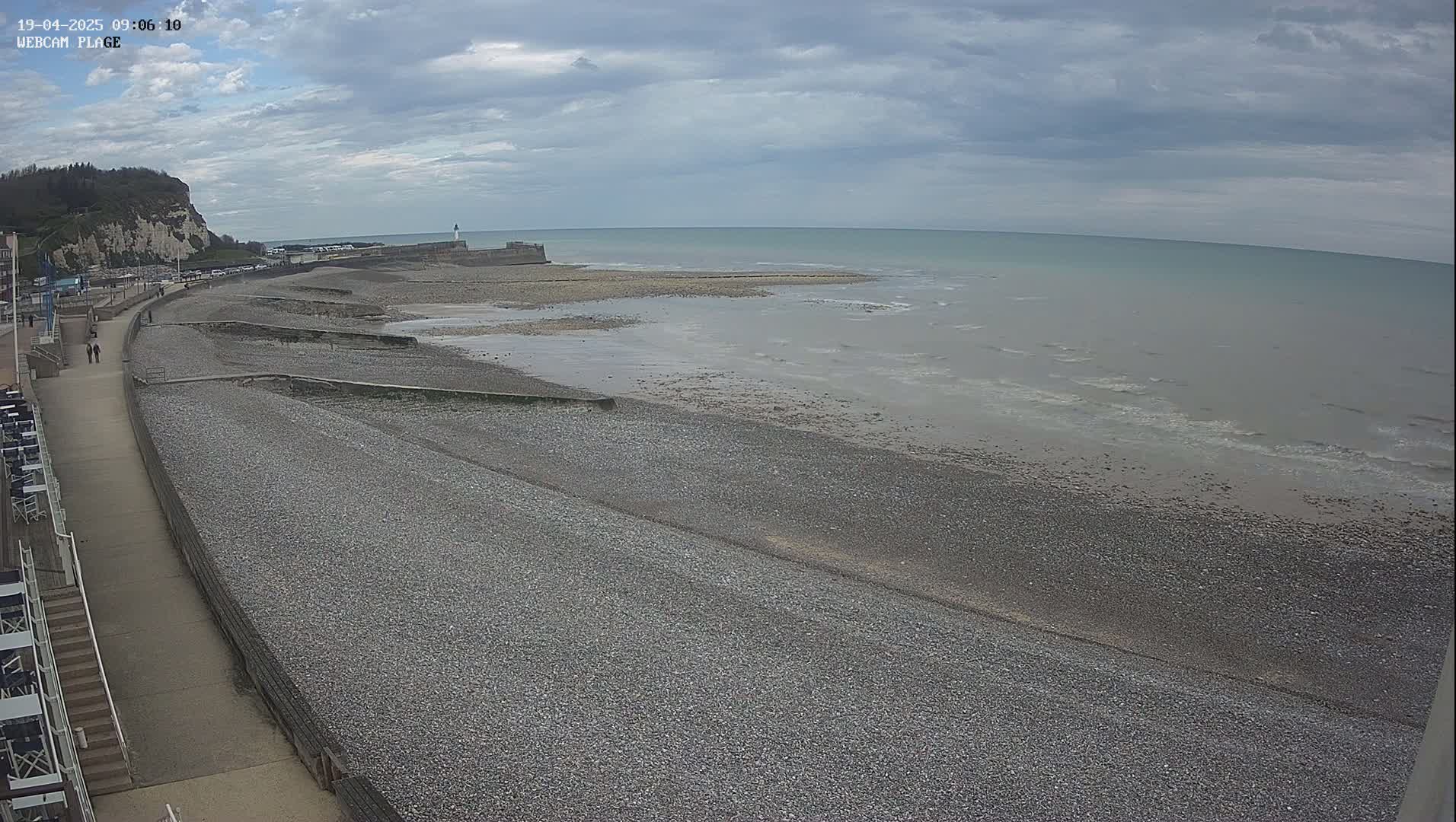 A pebble beach stretches alongside a paved walkway toward a calm sea under a cloudy sky, with a cliff and buildings visible in the distance.