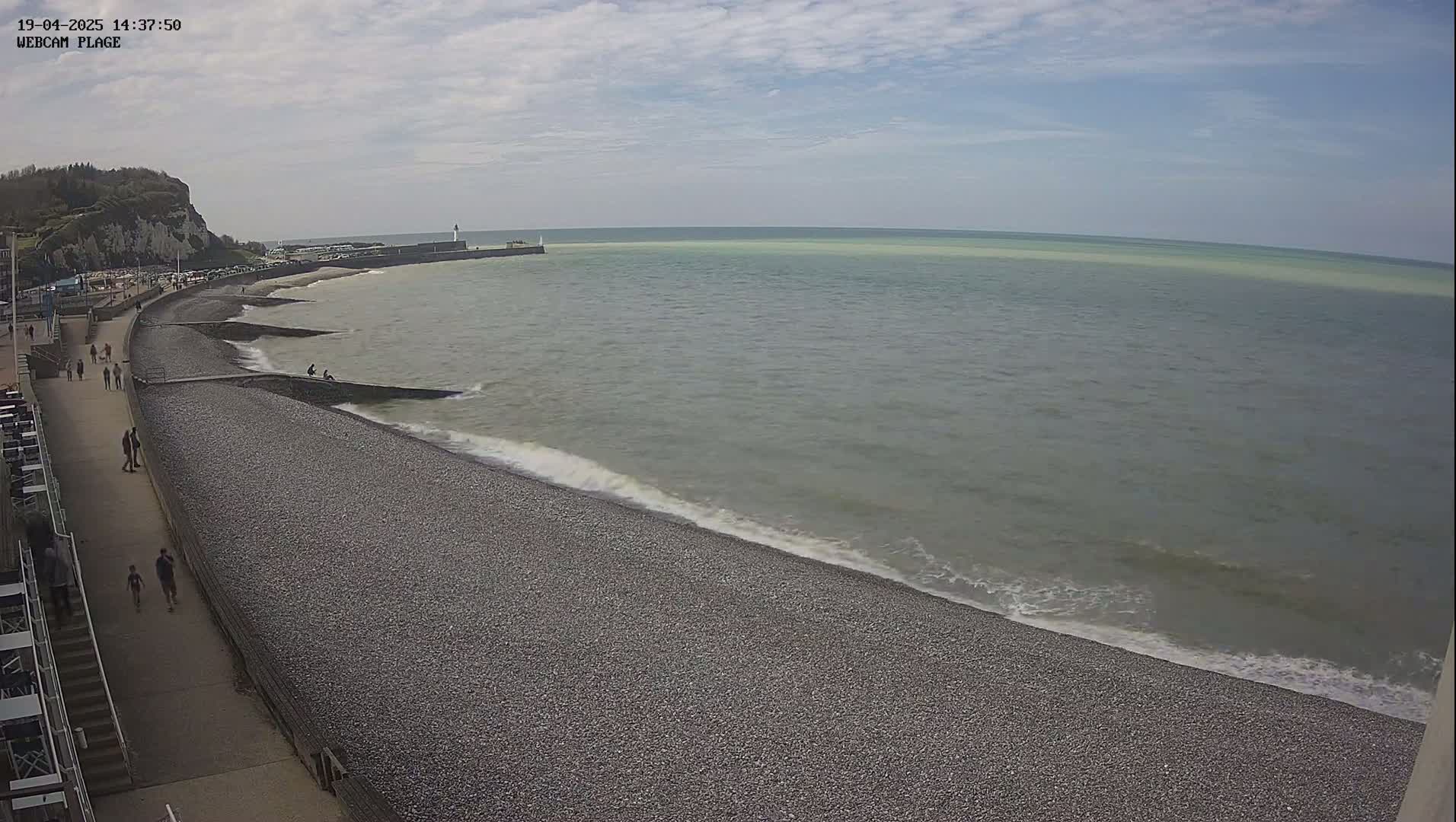 A pebble beach with a few people walking along a paved path, next to a calm sea with small waves under a partly cloudy sky.