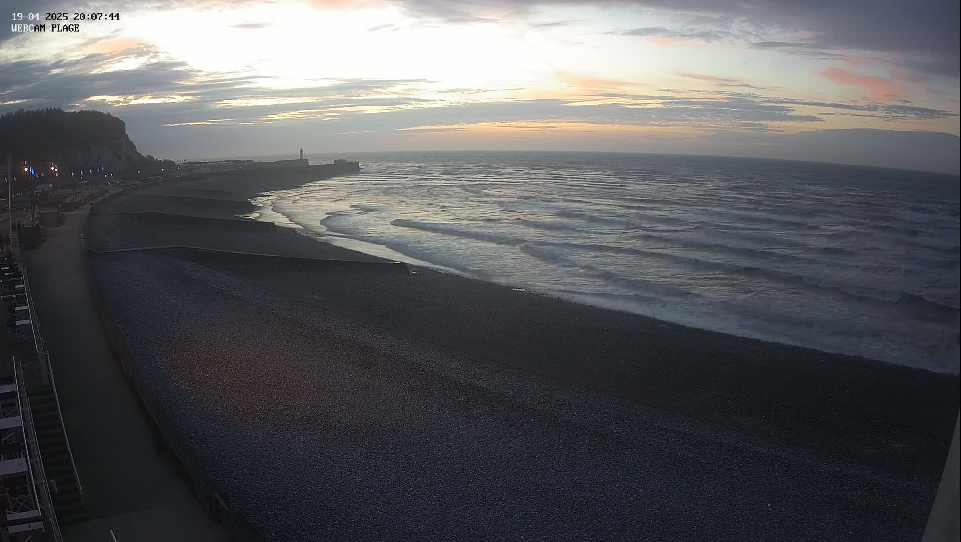 A dark pebble beach at sunset, with gentle waves rolling onto the shore under a partly cloudy sky.
