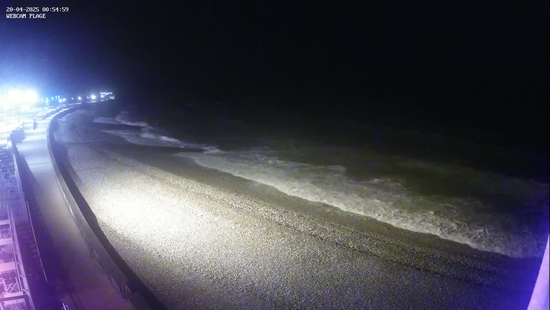 A dark, pebble beach at night is lit by artificial lights, with small waves lapping the shore.