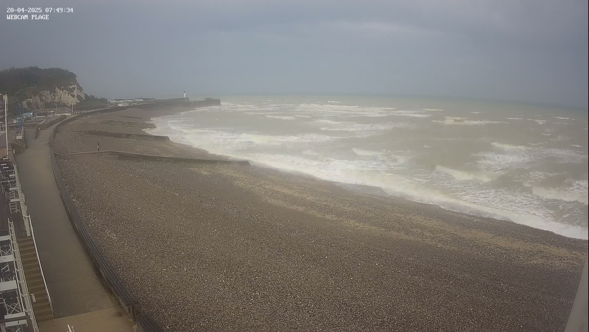 A pebble beach with a walkway and white cliffs in the background is seen under an overcast sky with choppy, gray waves rolling onto the shore.
