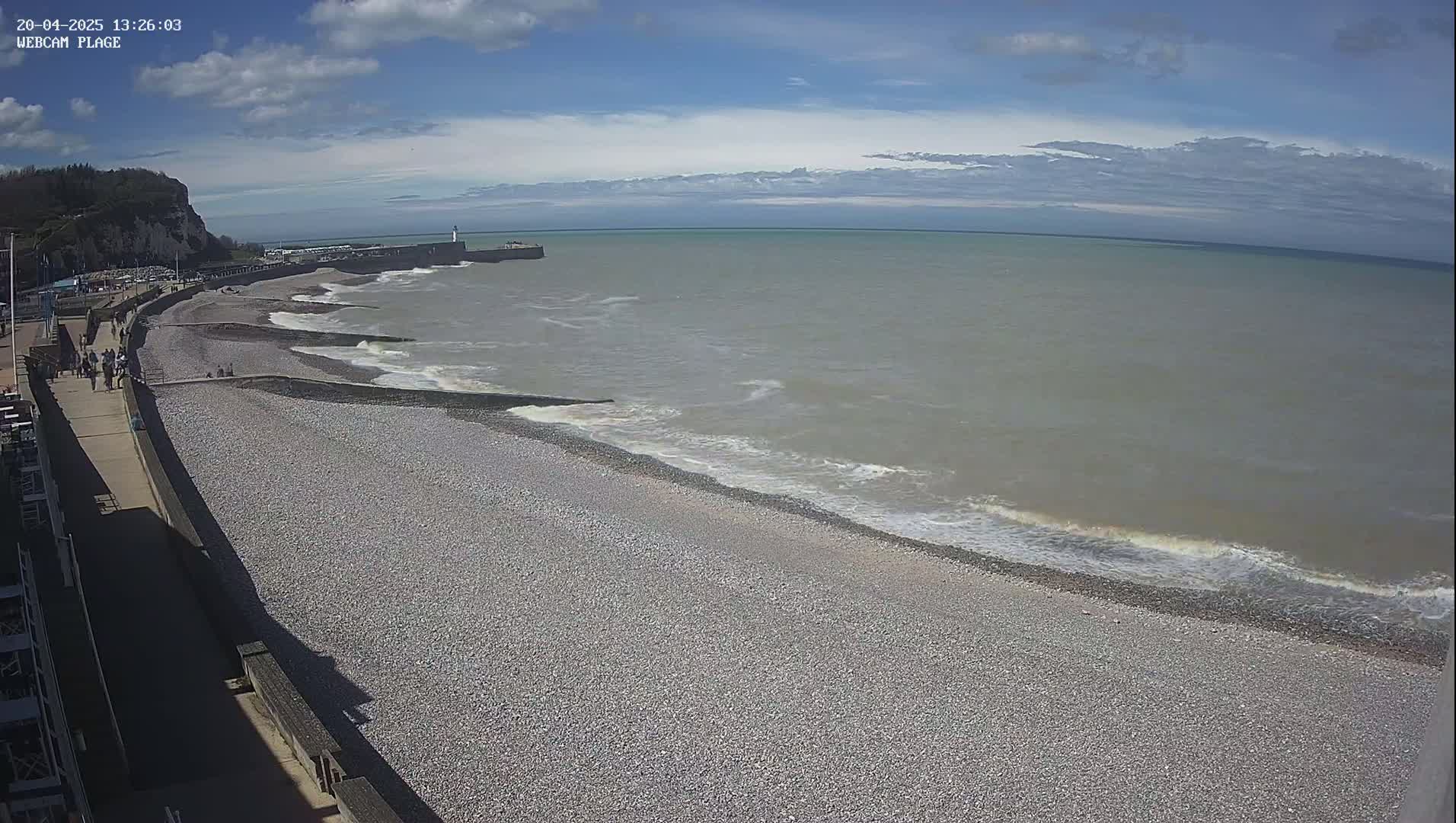 A pebble beach with people walking along a promenade next to a calm, grey sea under a partly cloudy sky.
