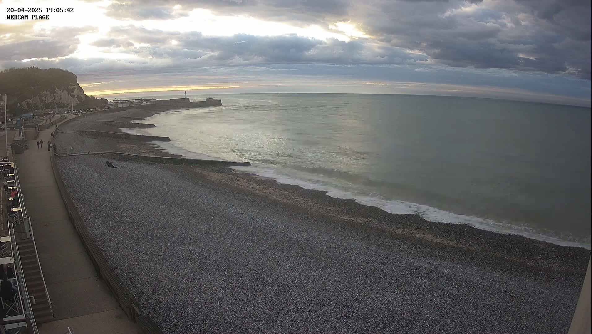 A pebble beach stretches along a calm sea under a partly cloudy sky, with a few people walking along the promenade.
