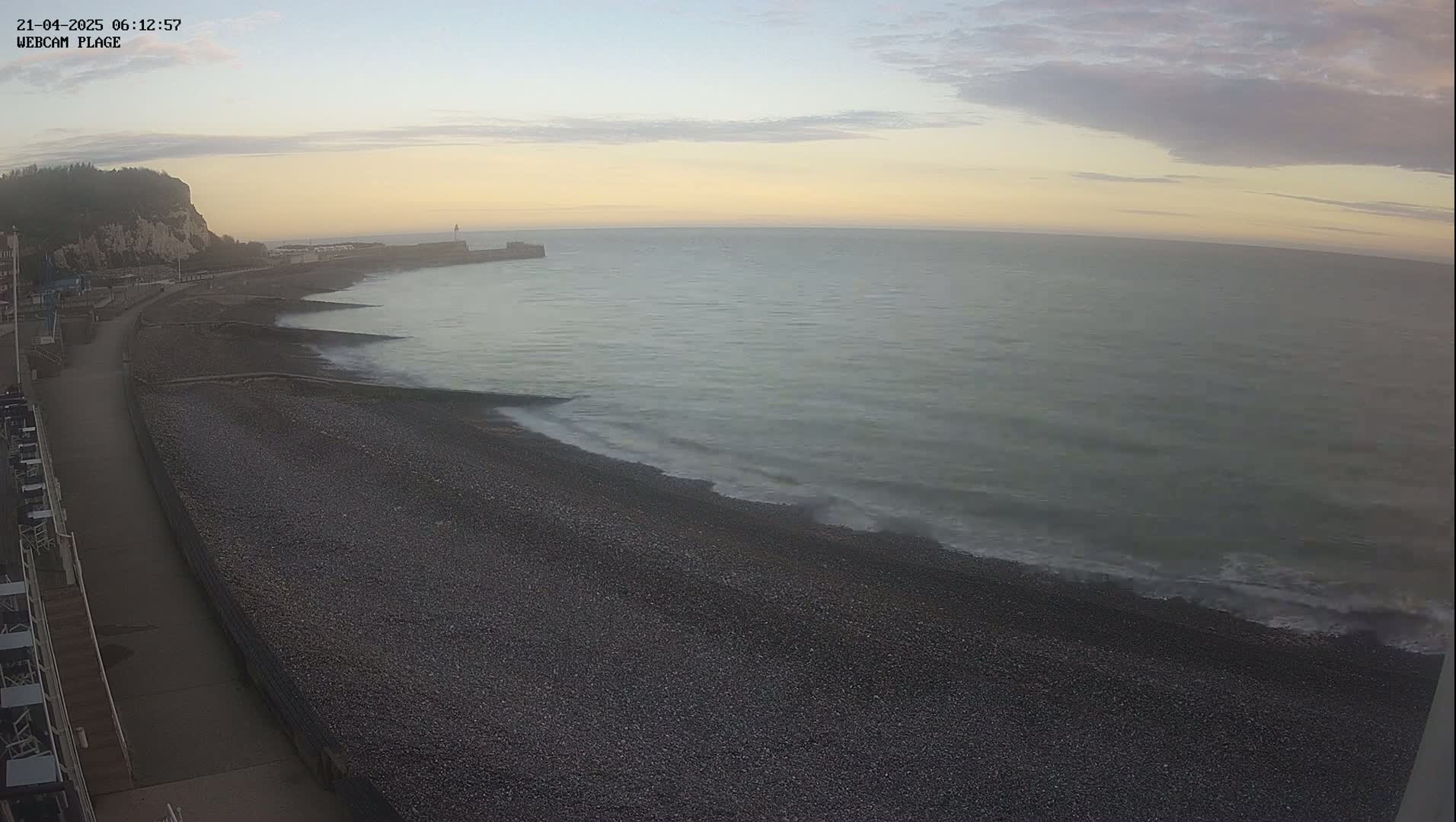 A pebble beach and calm sea are visible under an overcast sky at dawn, with a cliff and walkway in the background.