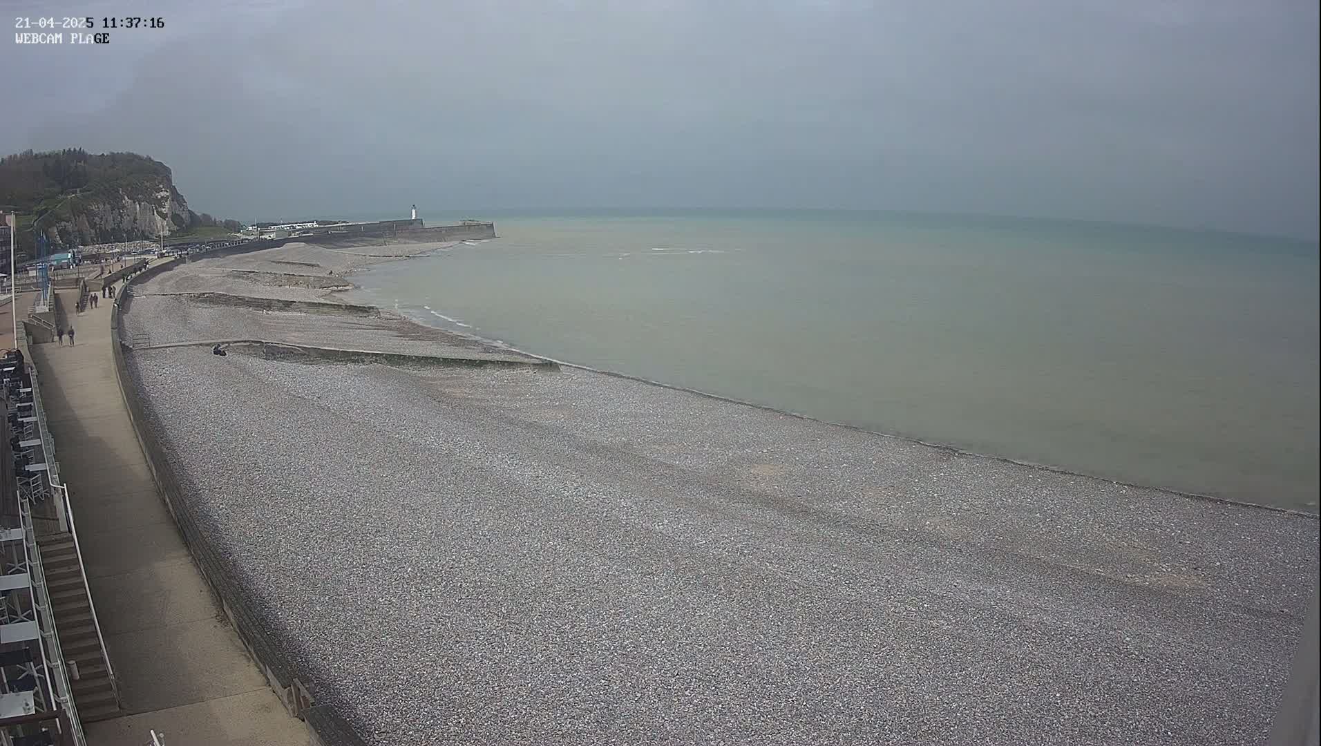 A pebble beach stretches out to a calm, gray-toned sea under an overcast sky, with a paved walkway and a few people visible along the shoreline.