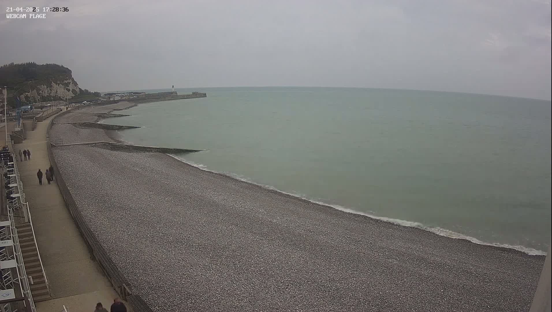 A pebble beach stretches along a calm, gray-green sea under an overcast sky, with a paved walkway and a few people walking along the shore.