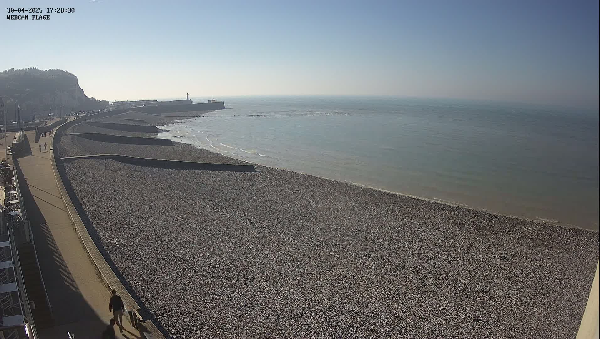A pebble beach stretches along a calm, light-blue ocean under a clear, sunny sky, with a paved walkway and a few people walking nearby.