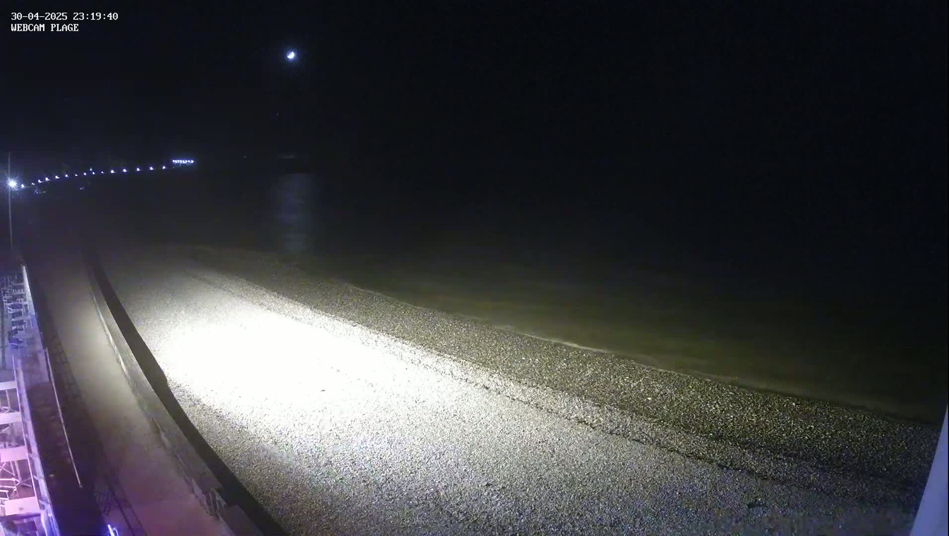 A pebble beach at night is illuminated by artificial light, with dark water visible in the background under a dark sky.